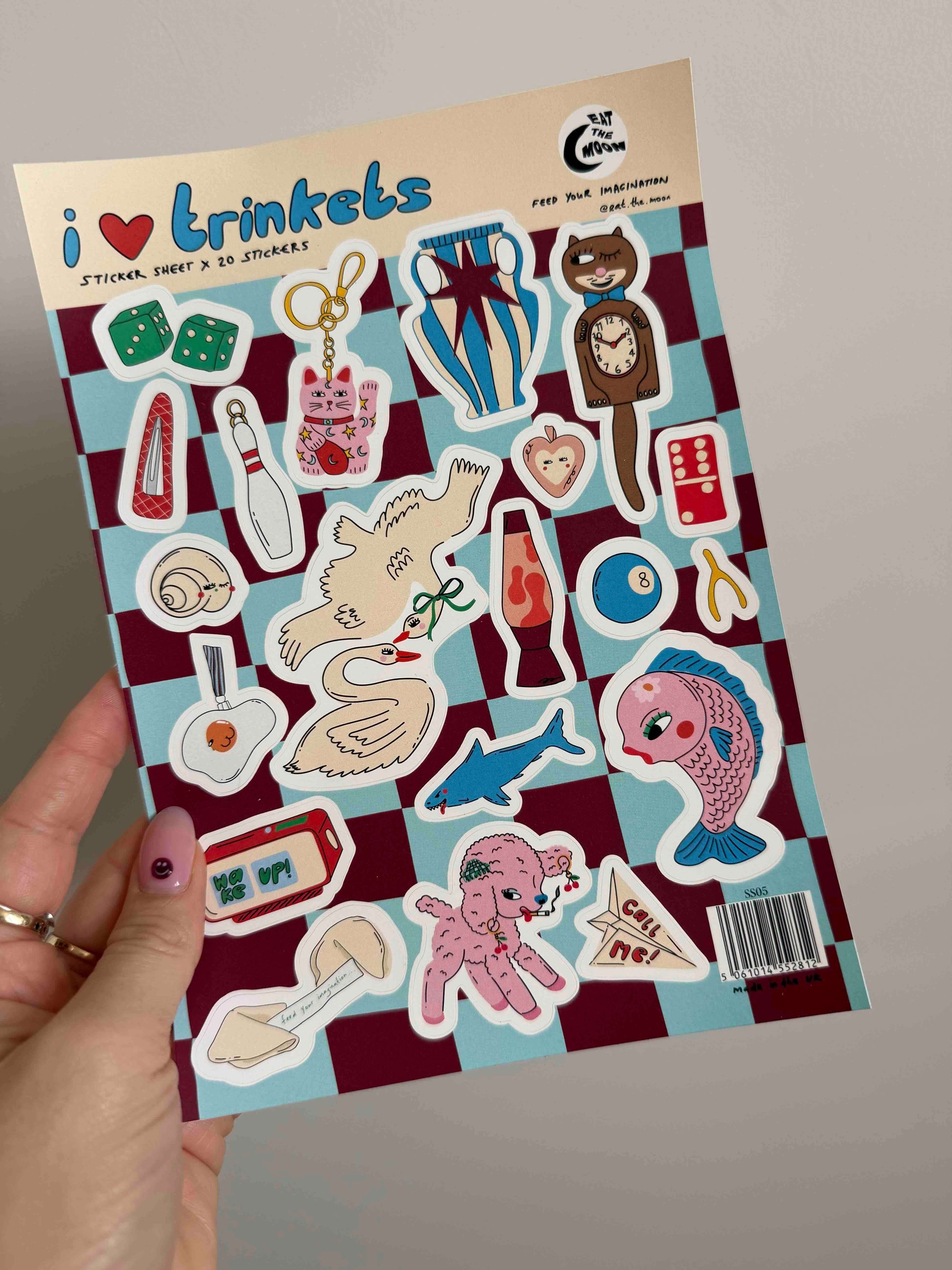 A hand holds a sticker sheet featuring various cartoon-style stickers on a maroon and light blue checkered background. The sheet includes illustrations of a cat, fish, snail, dice, bottle, and other objects.